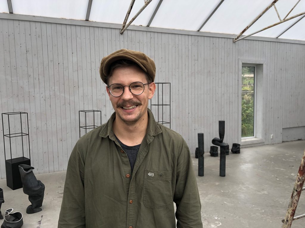 metalkonstnären Jonas Olsson står i ett vitt galleri med hans konstverk stående i bakgrunden